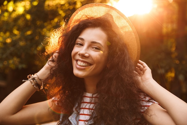 Foto close-up van brunette lachende vrouw met strooien hoed op zoek naar jou, tijdens een wandeling door park in zonnige dag
