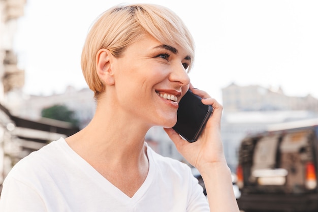 Foto close-up van lachende gelukkige vrouw dragen witte t-shirt zitten in zomerterras buiten, en met mobiel gesprek