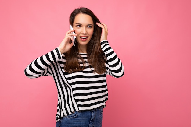 Foto die van aantrekkelijke emotionele jonge vrouw is ontsproten die op de smartphone spreekt die gestreepte sweater draagt