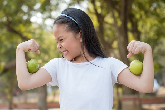 Foto gelukkig weinig aziatisch meisjeskind dat zich met grote glimlach bevindt. meisje met groene appel met biceps. verse gezonde groene bio-natuur met abstract wazig gebladerte en helder zomerzonlicht