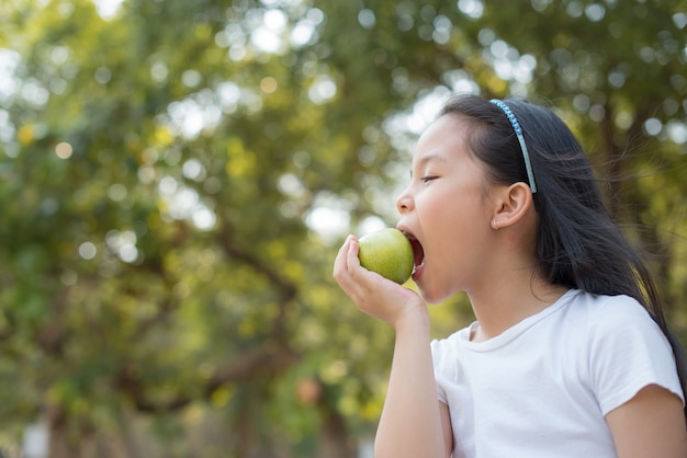 Foto gelukkig Weinig Aziatisch meisjeskind dat zich met grote glimlach bevindt. met groene appel in je hand verse gezonde groene bio-natuur met abstract wazig gebladerte en helder zomerzonlicht