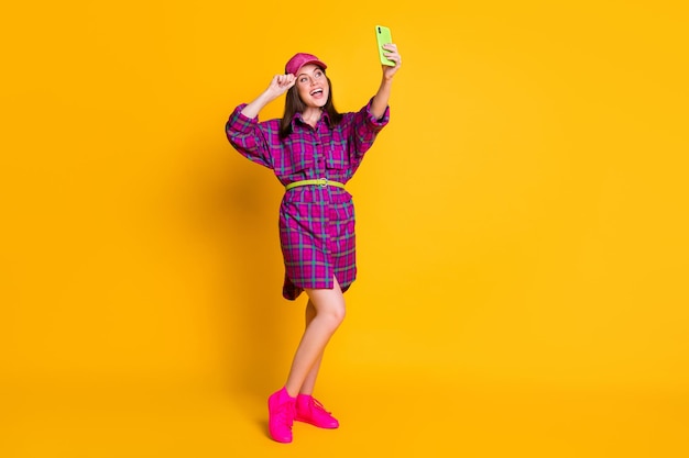 Foto op volledige grootte van een cool grappig tienermeisje dat selfie draagt, paarse geruite mini-jurkriem roze sneakers geïsoleerd op gele achtergrond