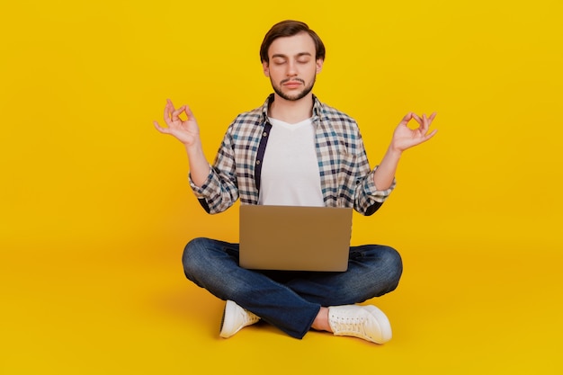 Foto op volledige grootte van een gelukkige jonge man die een laptop vasthoudt en yoga om mediteert terwijl hij op een vloer zit geïsoleerd op een gele achtergrond