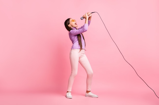 Foto op volledige grootte van een klein meisje dat een lied zingt op de microfoon, een paarse truibroek draagt, geïsoleerd op een pastelkleurige achtergrond