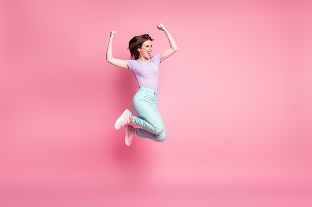 Foto op volledige grootte van opgetogen gek meisje springt geniet van loterij win hef vuisten op schreeuw draag turquoise violette broek sneakers geïsoleerd over pastelkleurige achtergrond