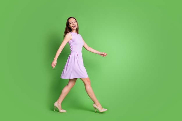 Foto op volledige grootte van schattige slanke jonge meisjesdame die vooruit loopt lege ruimte kijk camera brede glimlach advertentie kleding merk slijtage paarse jurk geïsoleerde pastel groene kleur achtergrond