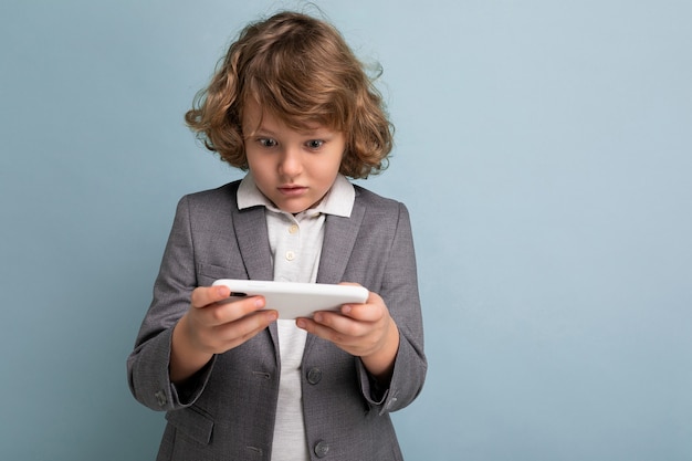 Foto-opname van een knappe emotionele kindjongen met krullend haar in een grijs pak dat een telefoon vasthoudt en gebruikt, geïsoleerd op een blauwe achtergrond, kijkend naar het spelen van smartphonespellen.