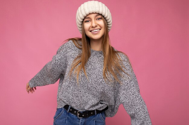 Foto-opname van een mooie glimlachende gelukkige jonge donkerblonde vrouw die geïsoleerd staat over een roze achtergrondmuur met een grijze trui en een beige winterhoed die naar de camera kijkt en plezier heeft.