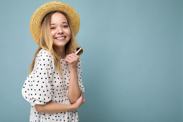 Foto-opname van een mooie positieve jonge blonde vrouw die zomerse vrijetijdskleding draagt en een stijlvolle zonnebril vasthoudt, geïsoleerd over een kleurrijke achtergrond die naar de camera kijkt