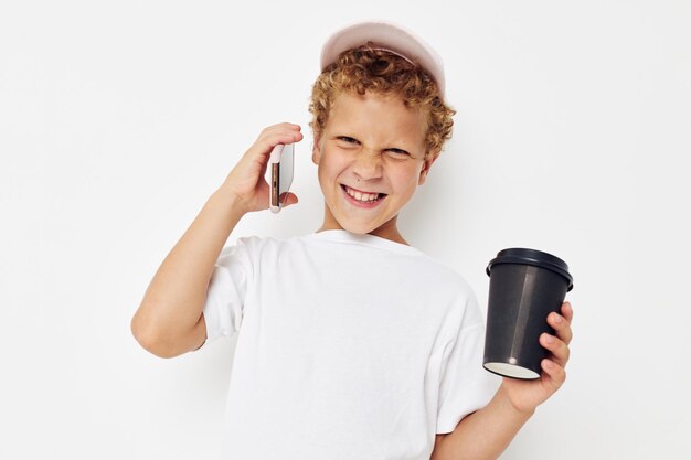 Foto portret krullend jongetje in een witte tshirt pet met een telefoon in een glas met een drankje lichte achtergrond ongewijzigd