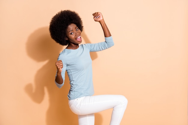 Foto portret van vrolijke afro-amerikaanse vrouw dansen met vuisten omhoog in de lucht geïsoleerd op pastel beige gekleurde achtergrond