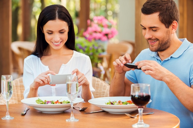 Foto's maken van eten. Mooie jonge verliefde koppels die foto's maken van hun eten en glimlachen terwijl ze samen in het openluchtrestaurant ontspannen