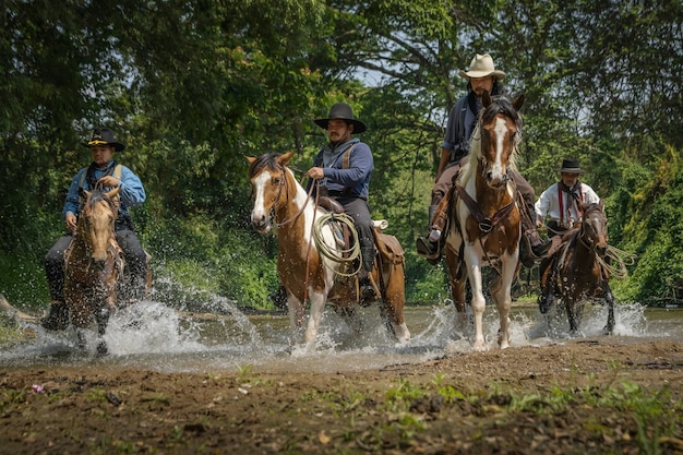 Foto's van veel mannen die cowboykleding dragen, op paarden rijden en over de rivier reizen