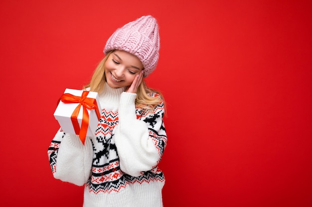 Foto schot van mooie gelukkig lachende verbaasde jonge blonde vrouw geïsoleerd over rode achtergrond muur dragen winter trui en roze hoed met witte geschenkdoos met rood lint. Ruimte kopiëren, mockup