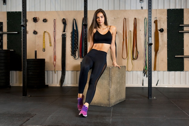 Foto training van een mooie brunette meisje in een zwarte sport uniform en roze sneakers die op een houten kubus in de sportschool zit