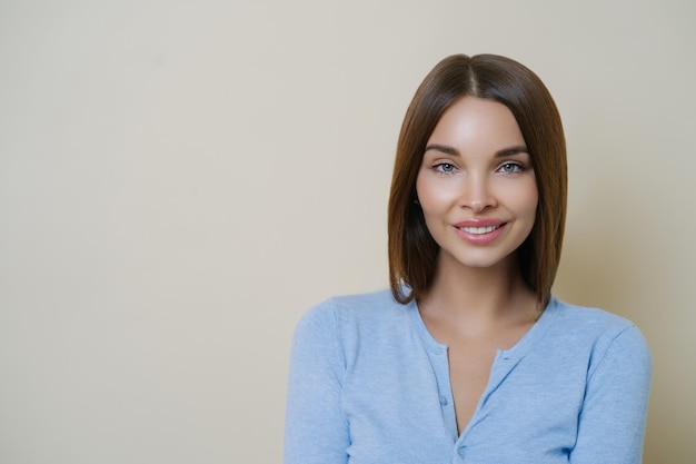 Foto van aanbiddelijke brunette vrouw heeft natuurlijke make-up en een gezonde, perfecte huid glimlacht teder toont witte tanden besteedt vrije tijd aan de zorg voor zichzelf draagt casual blauwe trui poses binnen