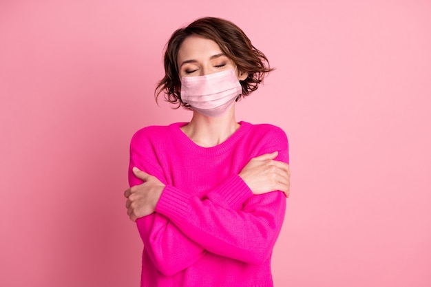 Foto van aantrekkelijke dame handen omhelzen zichzelf schouders genieten van vrolijke zachte doek wasgoed warmte ogen dicht draag casual heldere trui medisch masker geïsoleerd roze pastel kleur achtergrond