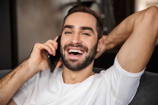 Foto van aantrekkelijke man 30s dragen casual t-shirt praten over smartphone zittend op de Bank in de woonkamer