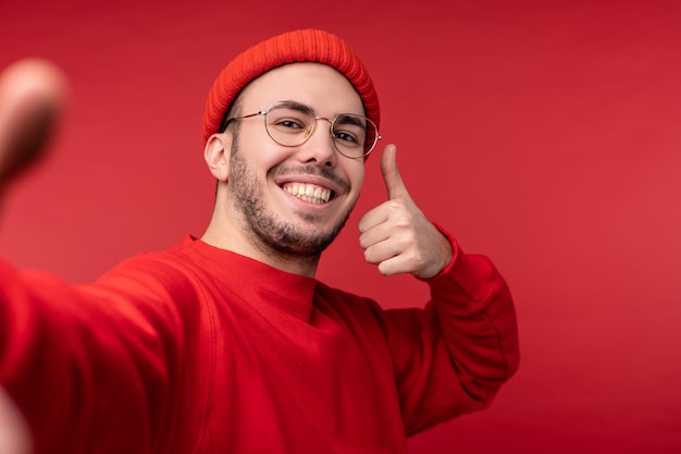 Foto van aantrekkelijke man met baard in glazen en rode kleding. Man geeft duim omhoog en neemt selfie en glimlacht, geïsoleerd op rode achtergrond.