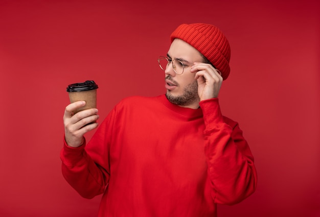 Foto van aantrekkelijke man met baard in glazen en rode kleding. Nieuwsgierige man kijkt naar zijn koffiekopje, geïsoleerd op rode achtergrond.