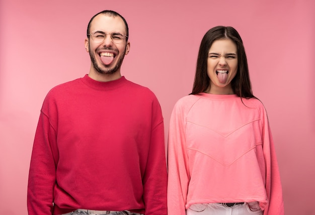 Foto van aantrekkelijke man met baard in rode kleding en vrouw in roze grimas met tong uitsteekt, geïsoleerd op roze achtergrond.