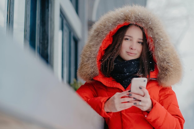 Foto van aantrekkelijke vrouw houdt moderne, moderne gadget vast, draagt rode jas met hoody, checkt e-mailbox aangesloten op draadloos internet, staat buiten geniet van vrije tijd en ijzig winterweer