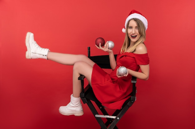 Foto van aantrekkelijke vrouw in kerstmuts en rode jurk, zit op een stoel, speelt met kerstspeelgoed en poseert met een glimlach. geïsoleerd op rode achtergrond.