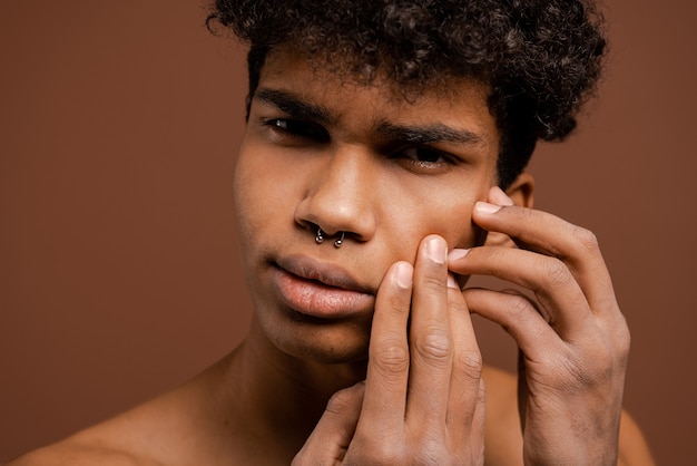 Foto van aantrekkelijke zwarte man met piercing raakt zijn gezicht om een puistje te laten knappen. Naakte torso, geïsoleerde bruine kleurenachtergrond.