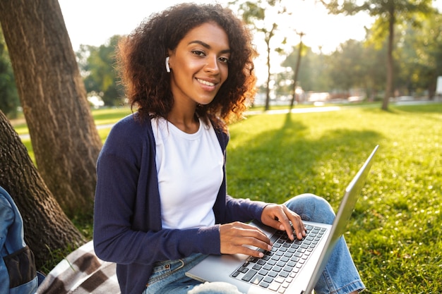 Foto van Afrikaanse jonge vrouw buiten zitten in park met laptopcomputer luisteren muziek met koptelefoon.