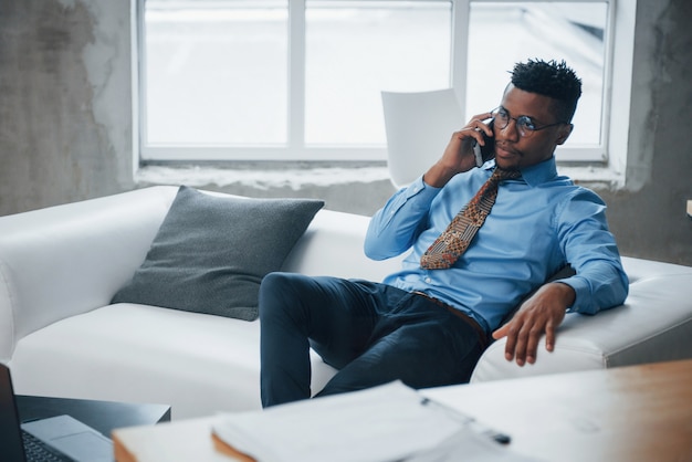 Foto van Afro-Amerikaanse jonge man zittend op een bank en gesprek op telefoon