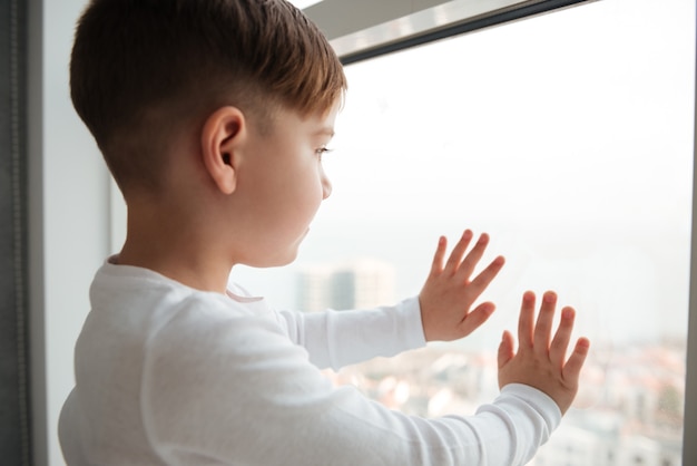Foto van alleen kleine jongen in de buurt van raam wachtend op ouders thuis. Kijk naar raam.