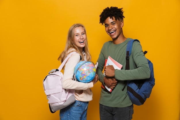 Foto van Amerikaanse en blanke studenten, man en vrouw 16-18 die rugzakken dragen met een aardbol, geïsoleerd op gele achtergrond yellow