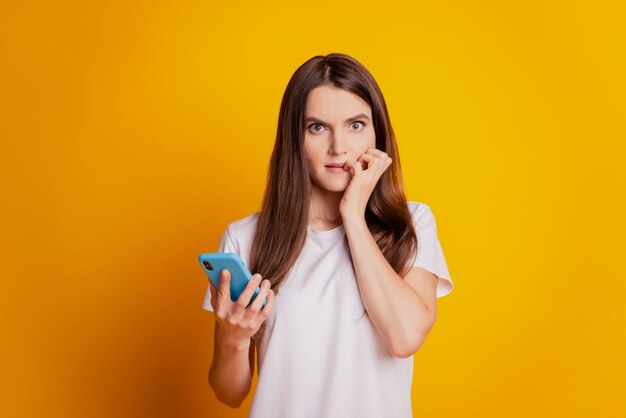 Foto van bezorgde dame die nagels bijt, telefoon vasthoudt, wit t-shirt draagt dat op gele achtergrond staat