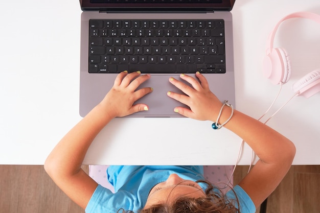 Foto van bovenaf van krullend donkerbruin meisje met handen op laptop bovenop witte bureautafel