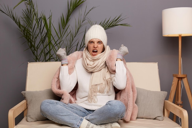 Foto van boze wanhoop vrouw met pet en winterjas zittend op de bank thuis schreeuwend met opgeheven vuisten en woede voelt zich depressief centrale verwarming werkt niet