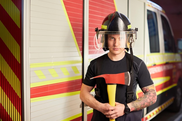 Foto van brandweerman met helm met bijl tegen brandweerwagen Afbeelding van jonge brandweerman met helm in hoofd tegen de achtergrond van brandweerwagen