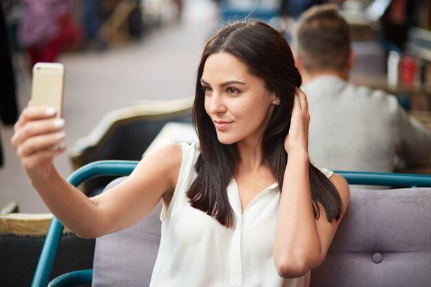 Foto van brunette jonge dame neemt selfie met moderne mobiele telefoon, gekleed in witte blouse, maakt foto- of videogesprek, geniet van haar uiterlijk