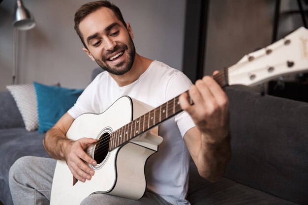 Foto van brunette man 30s dragen casual t-shirt akoestische gitaar spelen zittend op de Bank in appartement