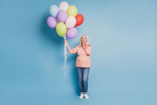 Foto van charmante oude vrouw die luchtballonnen vasthoudt in roze trui op blauwe achtergrond