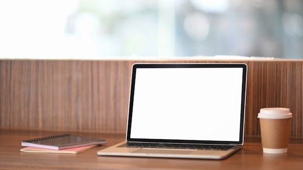 Foto van computerlaptop met het witte lege scherm die op houten lijst met koffiekop en notitieboekje zetten over ordelijke woonkamer.