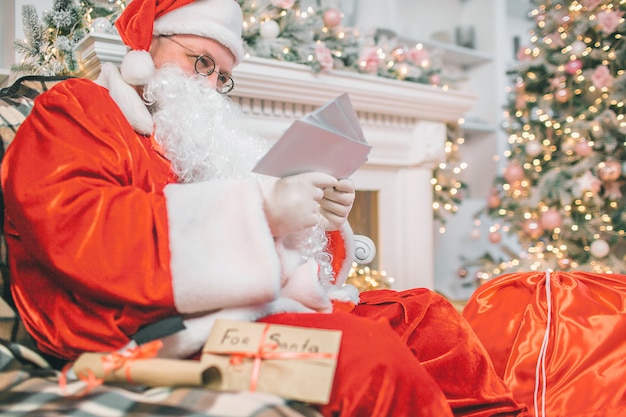 Foto van de kerstman zitten en lezen van brieven. Hij houdt ze in handen. De mens is geconcentreerd en serieus.