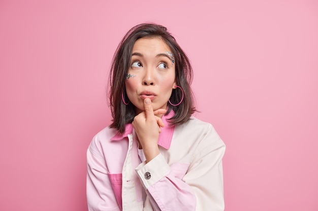 Foto van doordachte dromerige vrouw met oosterse uitstraling kijkt omhoog houdt duim in de buurt van lippen make-up haar mins probeert te beslissen over iets gekleed in modieuze kledingmodellen binnen tegen roze muur