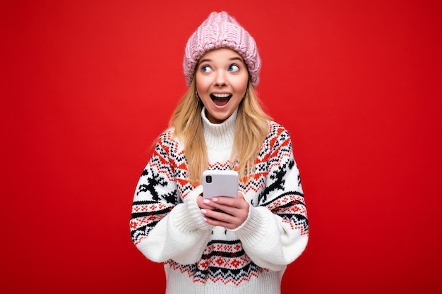 Foto van een aantrekkelijke, gekke verbaasde jonge vrouw die casual stijlvolle kleding draagt die geïsoleerd staat over de achtergrond met kopieerruimte die een mobiele telefoon vasthoudt en naar de zijkant kijkt.