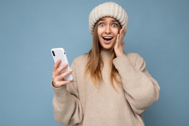 Foto van een aantrekkelijke, gekke verbaasde verraste jonge vrouw die casual stijlvolle kleding draagt die geïsoleerd staat over de achtergrond met kopieerruimte die een mobiele telefoon vasthoudt en naar de camera kijkt