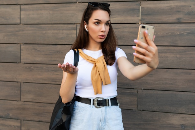 Foto van een aantrekkelijke, geschokte, verraste jonge vrouw met vrijetijdskleding die op straat staat te praten op de mobiele telefoon en naar smartphone kijkt.