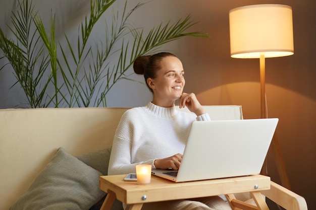 Foto van een aantrekkelijke glimlachende positieve optimistische vrouw die een witte trui draagt die op de bank zit en op een laptop werkt die wegkijkt en droomt over het einde van de werkdag