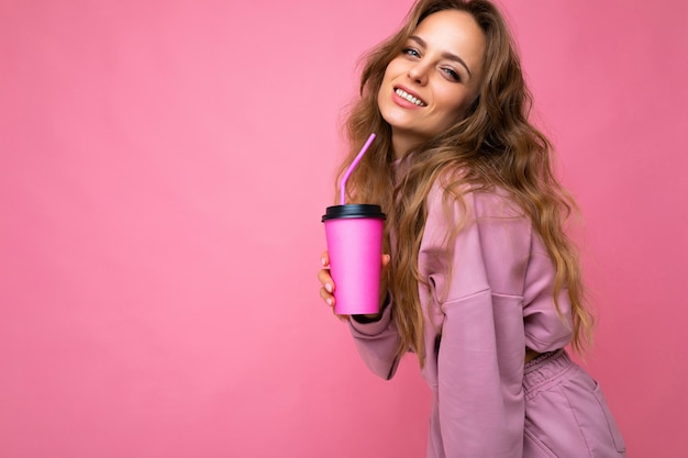 Foto van een aantrekkelijke jonge gelukkig lachende brunette vrouw die alledaagse stijlvolle kleding draagt, geïsoleerd over een kleurrijke achtergrondmuur met een papieren beker voor uitgesneden thee die naar de camera kijkt. kopieer ruimte