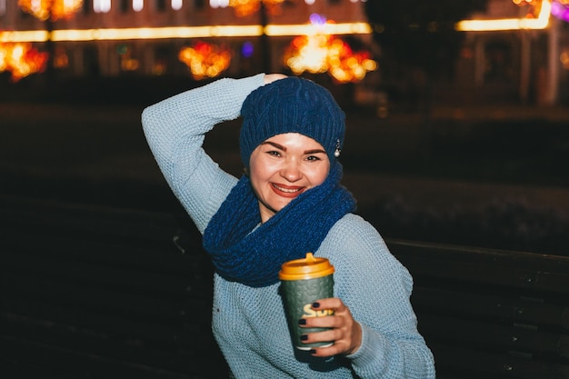 Foto van een aantrekkelijke, vrolijke vrouw die bij koud sneeuwweer op een bank zit, glimlacht en buiten koffie drinkt