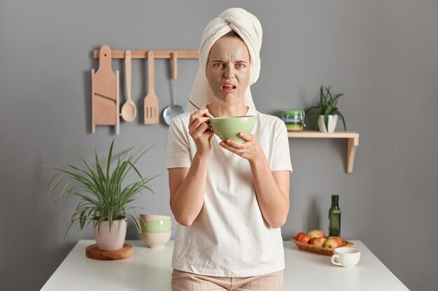 Foto van een bange blanke vrouw met een handdoek op het hoofd die sprakeloos binnen staat met een schoonheidsgezichtsmasker om fijne lijntjes te verminderen eet haar ontbijt wordt vroeg in de ochtend wakker poserend in de keuken