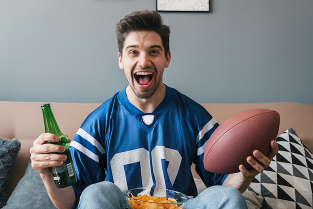 Foto van een blanke opgetogen man met een rugbybal die schreeuwt en bier drinkt terwijl hij naar een sportwedstrijd in de woonkamer kijkt